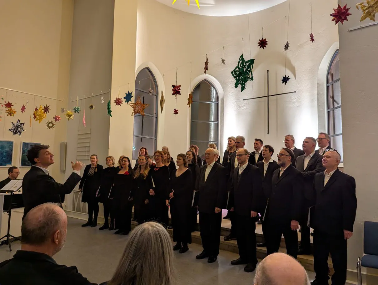 Chor steht in Choraufstellung in schwarzer Kleidung in der Friedenskirche. Der Chorleiter gibt die Töne und der Chor ist bereit zum Singen.